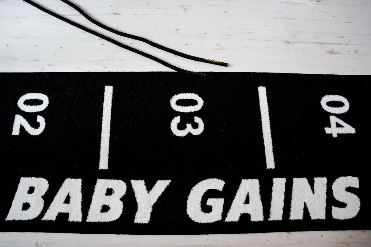 Top-down view of the black Baby Gains Grass Mat with white BABY GAINS lettering and 02, 03, 04 lane markers