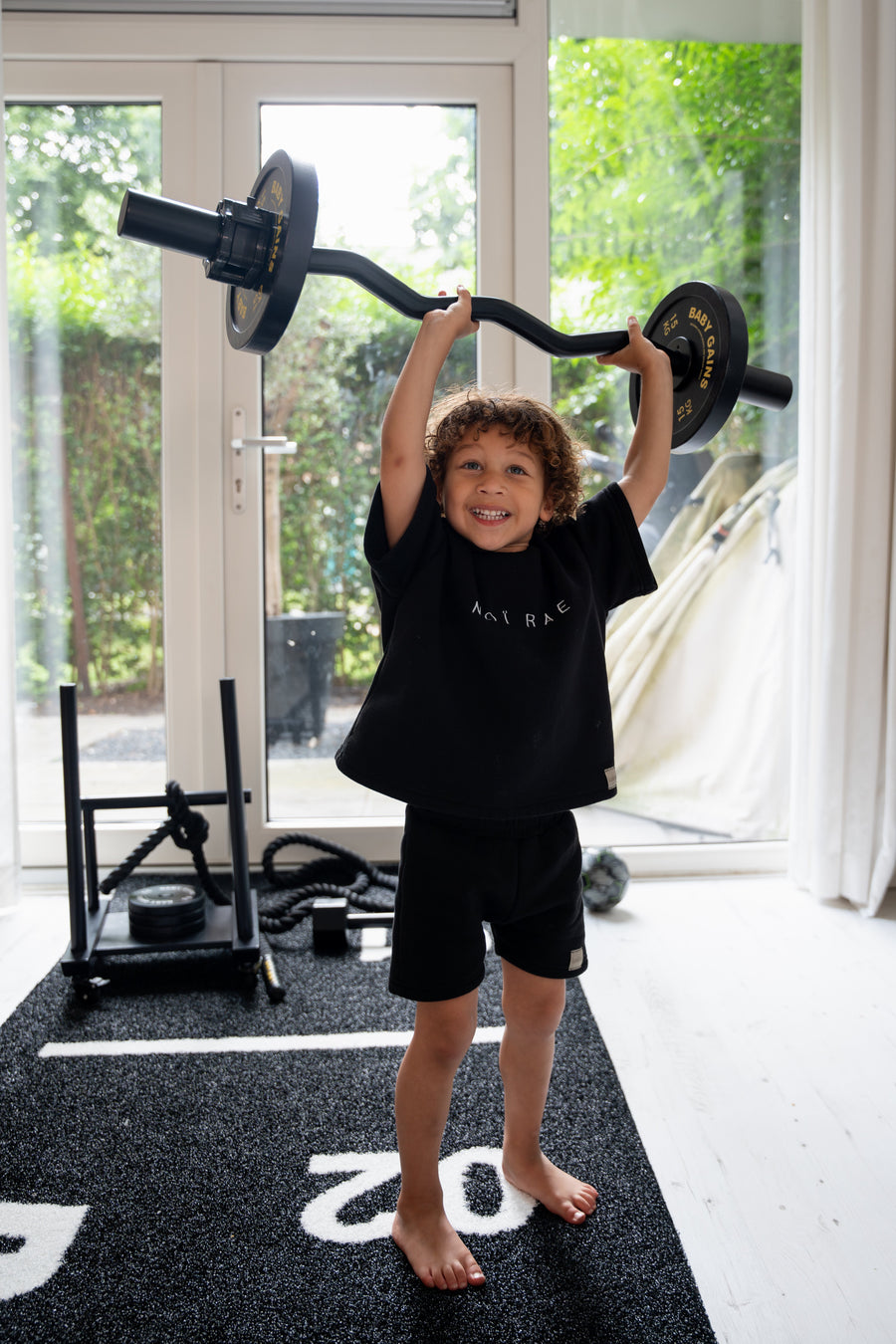 Barefoot boy in black lifting the loaded Kids Curl Bar overhead on a gym mat in front of garden doors