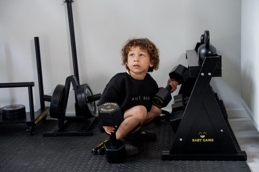 Curly-haired boy crouching with a Baby Gains hex dumbbell in each hand next to the Kids Elite Dumbbell Rack