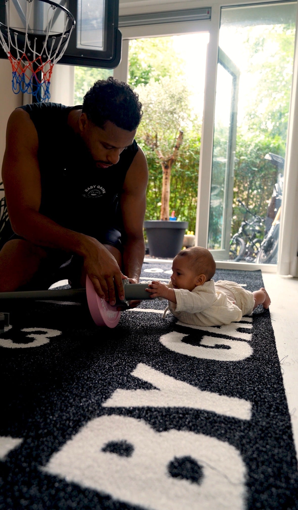 Dad loading a pink plate onto a kids barbell on the Baby Gains Grass Mat while his baby crawls toward him