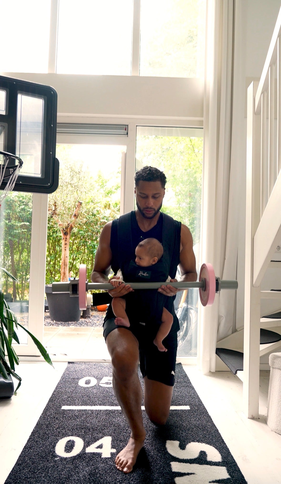 Dad lunging with the Kids Pink Starter Barbell while carrying a baby in a chest carrier on a BabyGains mat at home