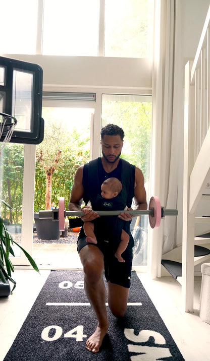 Dad lunging with the Kids Pink Starter Barbell while carrying a baby in a chest carrier on a BabyGains mat at home