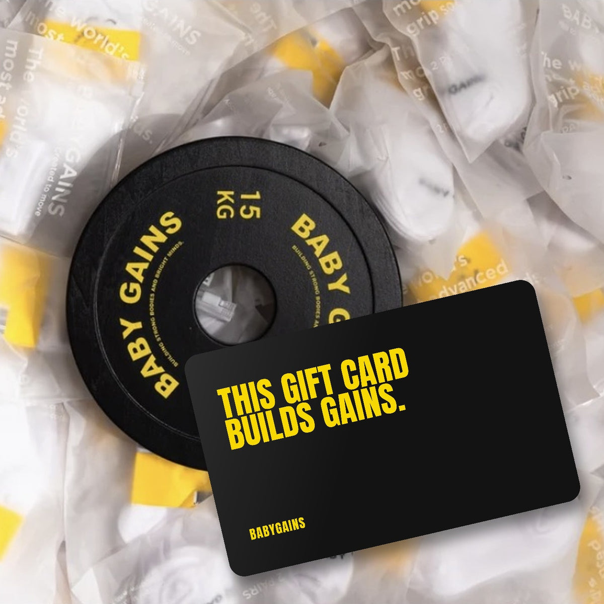 BabyGains gift card resting beside a black 15 kg BabyGains weight plate on a bed of packaged kids grip socks