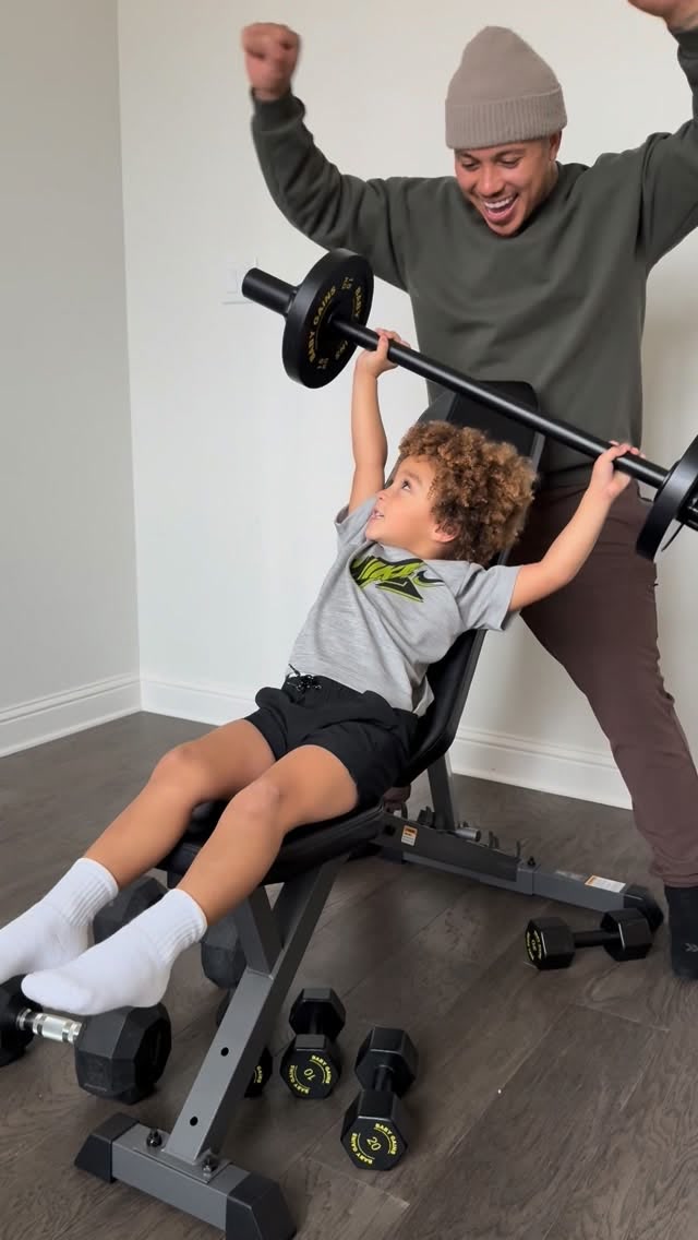A dad cheers while his son presses a Baby Gains barbell on a bench